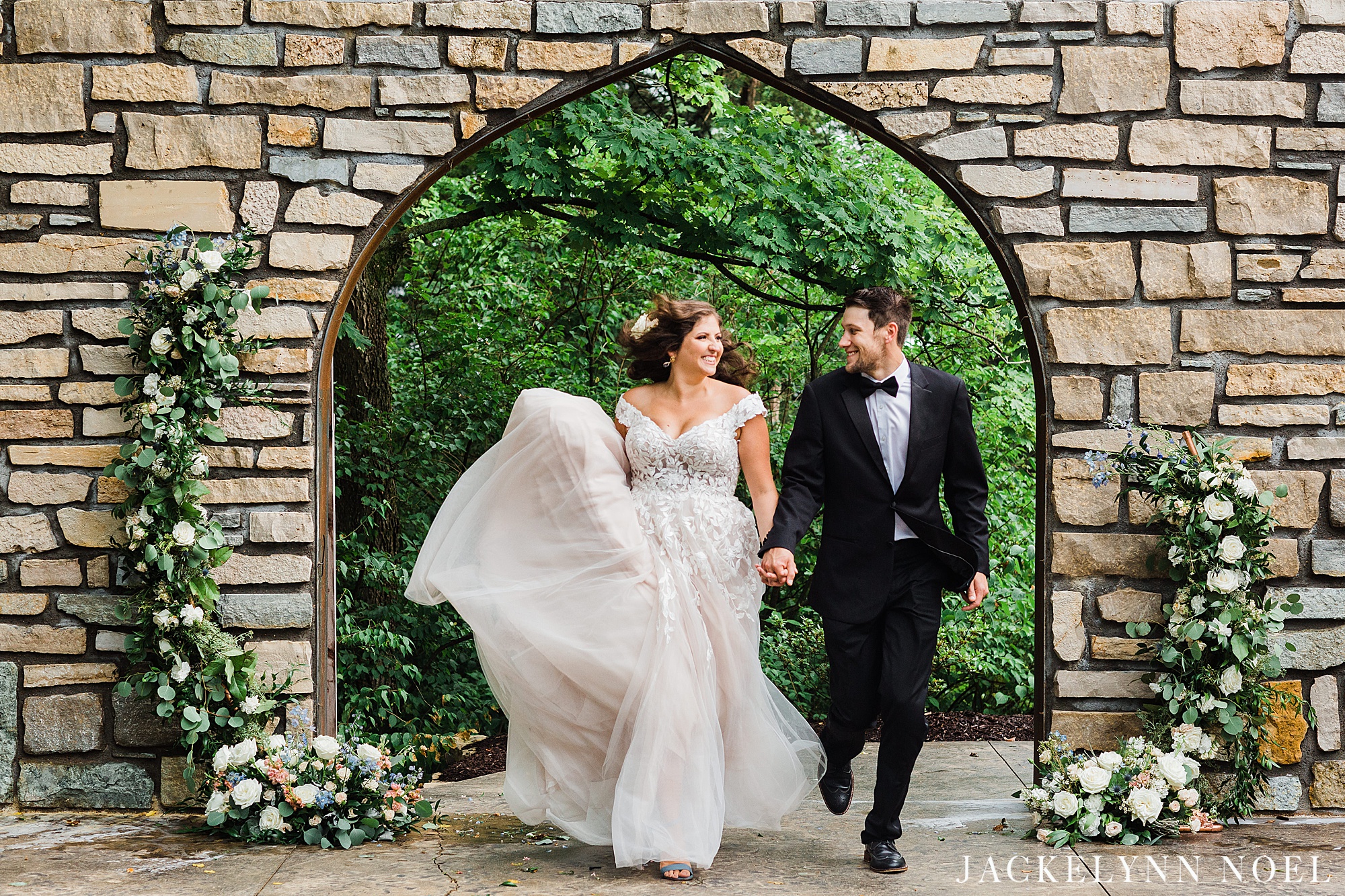 Bride and groom running through stone arch. Bride is wearing an off the shoulder dress and groom is in a classic black tuxedo.