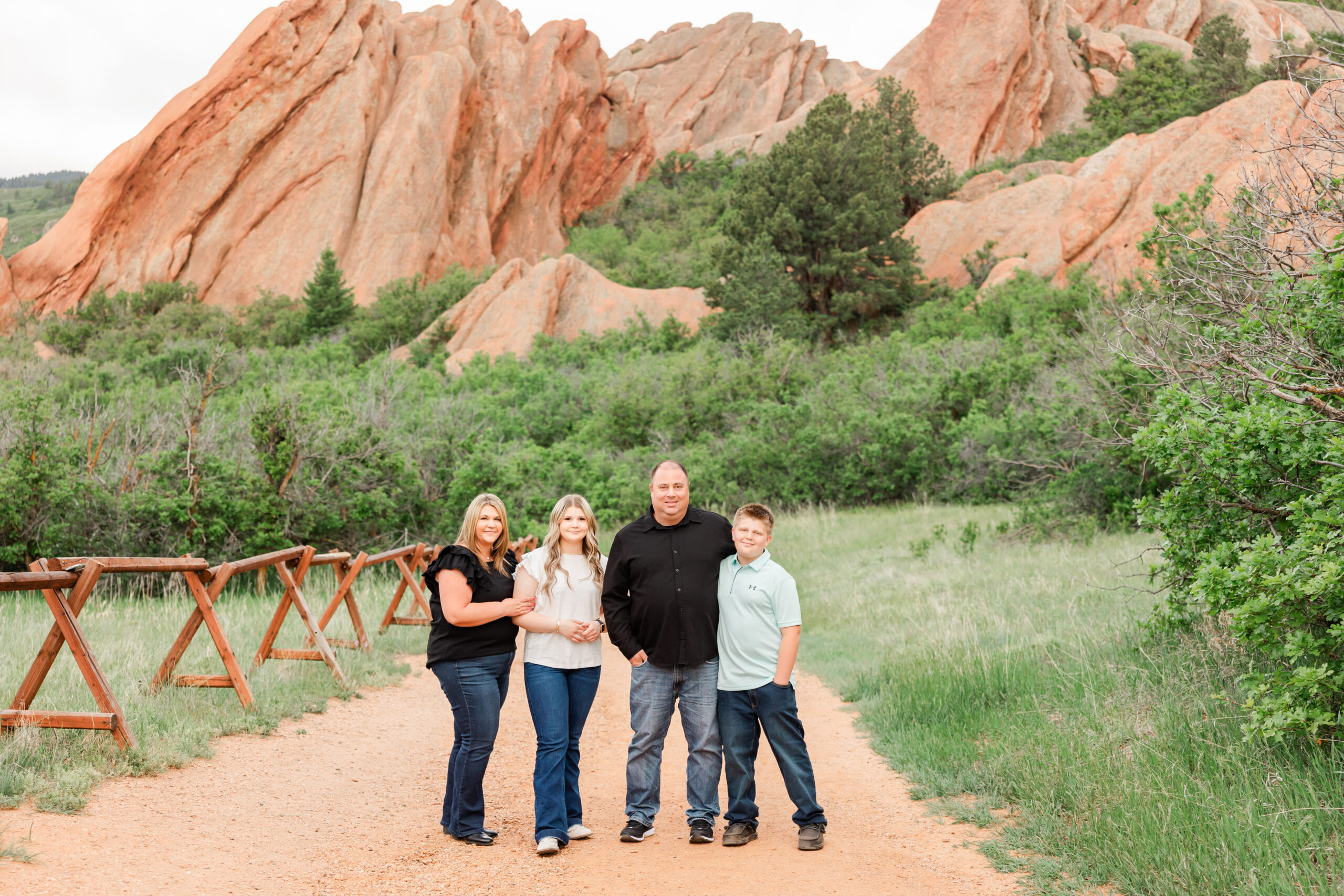 A family smiling together during a portrait session at Roxborough State Park, surrounded by massive, angled red rock formations and green foliage under a clear sky.